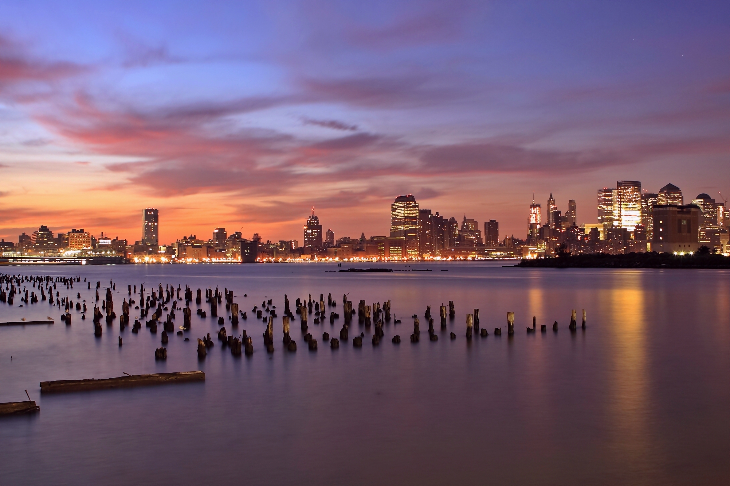 cities, rivers, sunset, sky, lilac, clouds, usa, orange, lights, wooden, skyscrapers, backlight, illumination, evening, united states, pillars, jersey city, state of new jersey, new jersey state, hudson, columns